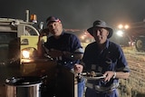 Two harvest workers holding plates and eating in the paddock at night with equipment in the background.
