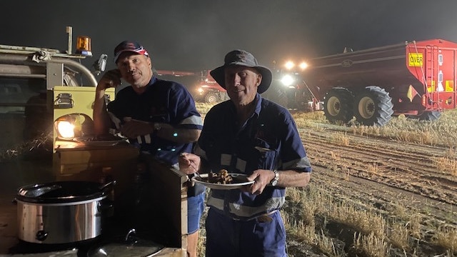 Two harvest workers holding plates and eating in the paddock at night with equipment in the background.