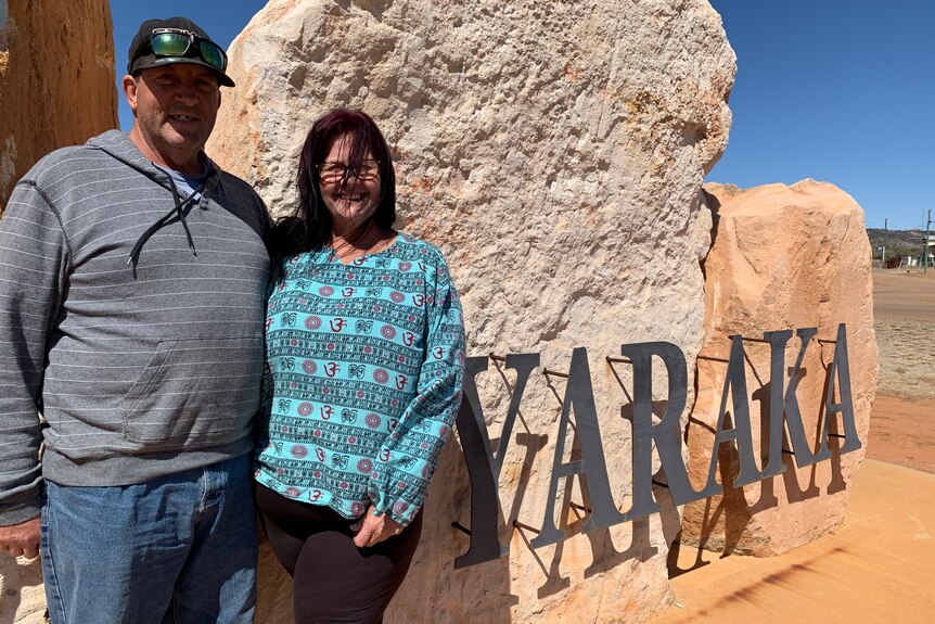 A man in a grey jumper and a lady in a blue top stand in front a big rock, with the word Yaraka made of steel attached to it.