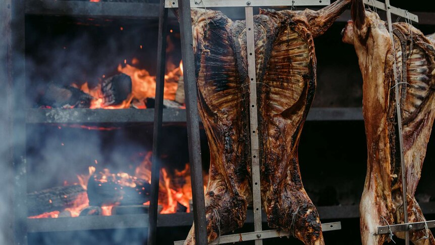 Picture of two whole cooked pigs hanging outdoors with fire in background.