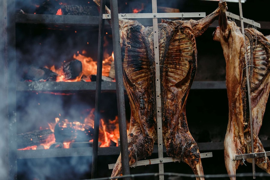 Picture of two whole cooked pigs hanging outdoors with fire in background.