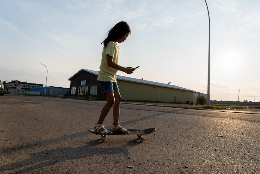A silhouette of a young girl skating on an empty street in the late afternoon sun. She looks down at an iPhone in her hands.