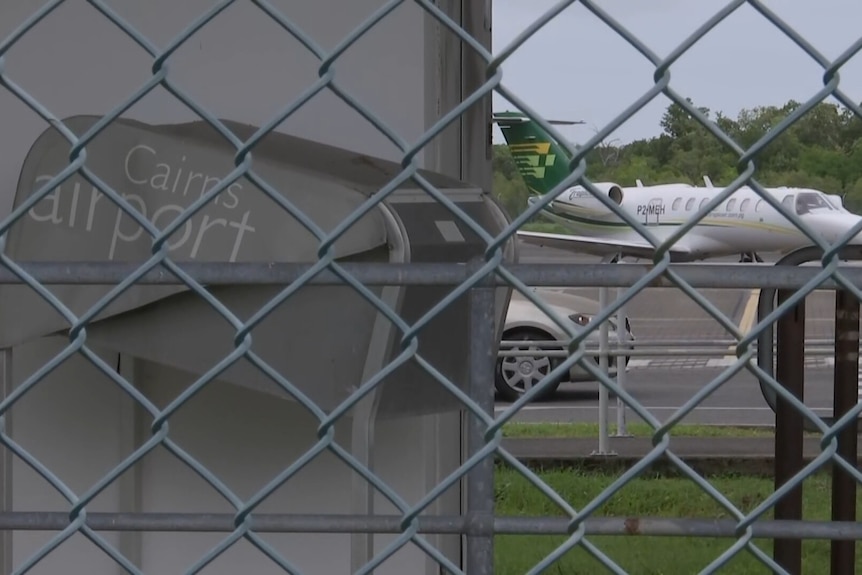 A small plane on the tarmac at an airport, as seen through a fence bearing a sign that reads "Cairns Airport".