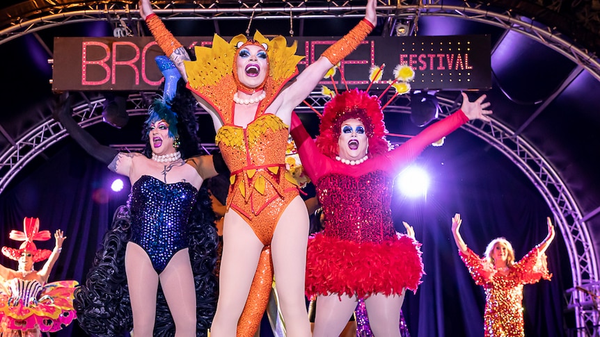 Three drag queens wearing colourful dresses and leotards dance up on stage.