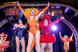 Three drag queens wearing colourful dresses and leotards dance up on stage.