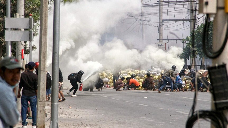 Protesters wearing helmets duck for cover behind a smoke bomb on an Asian street.