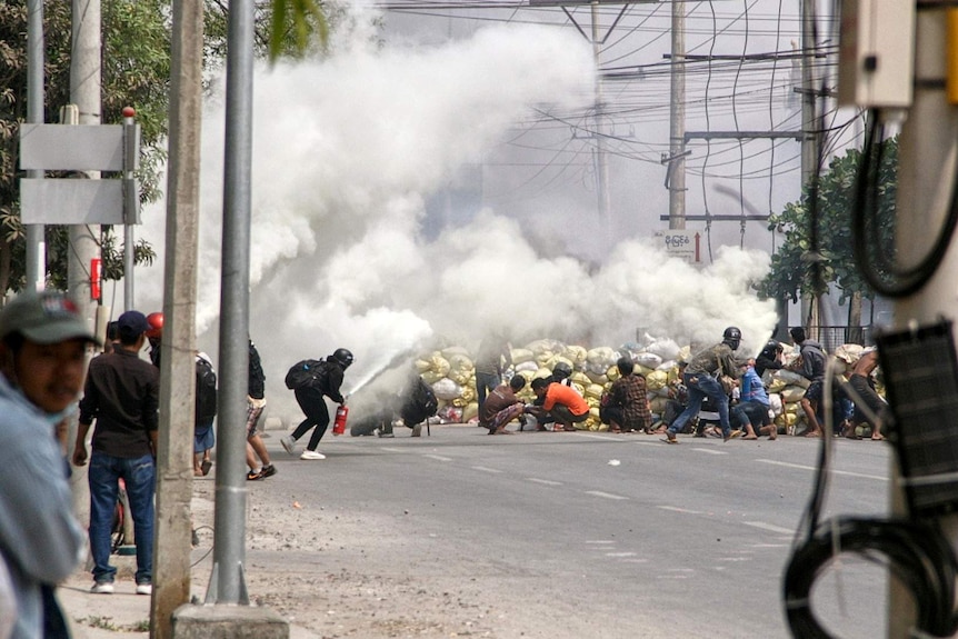 Protesters wearing helmets duck for cover behind a smoke bomb on an Asian street.