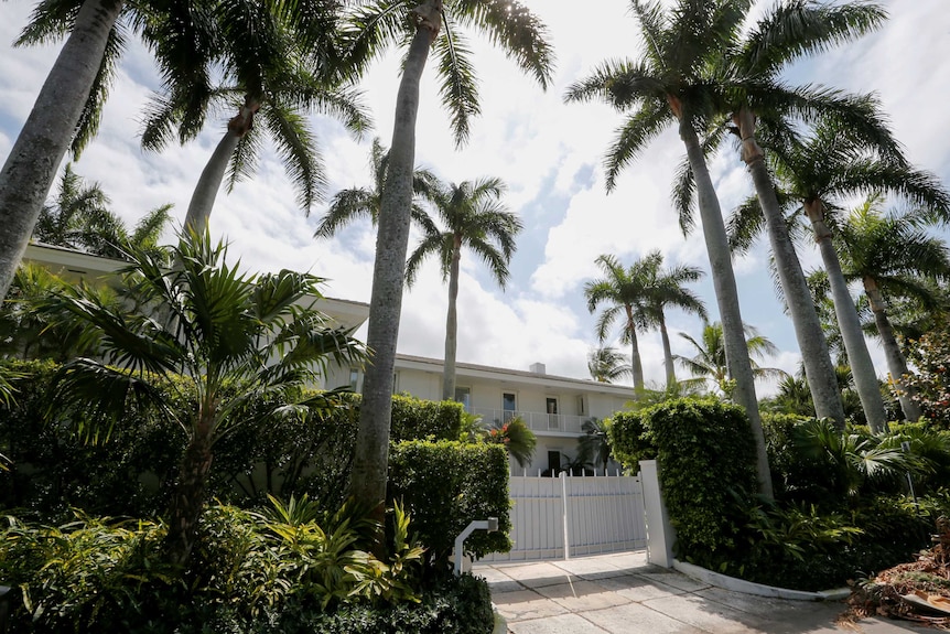 A white gate blocks access to a sprawling white home, with lush gardens and towering palm trees in front.