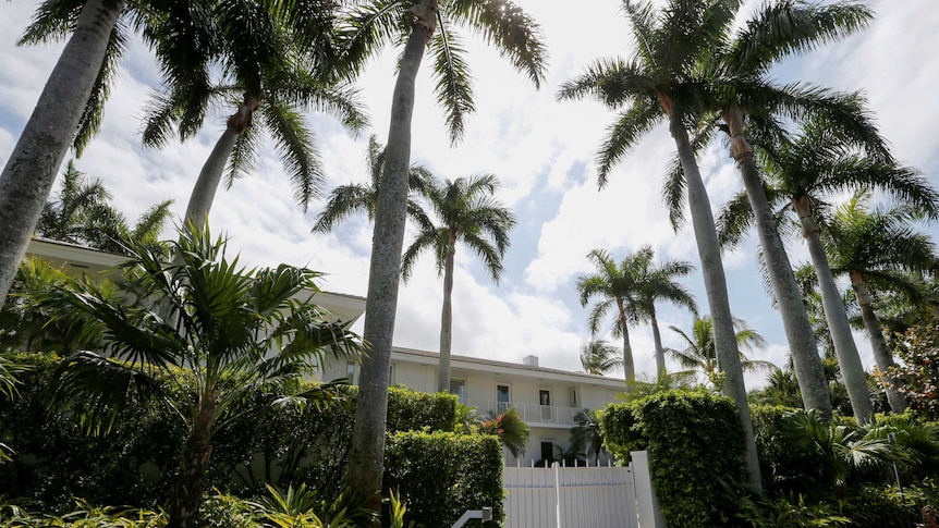 A white gate blocks access to a sprawling white home, with lush gardens and towering palm trees in front.