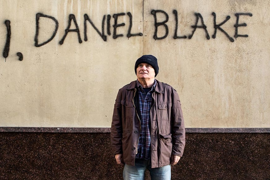 A man standing on a street with I, Daniel Blake painted on wall behind him, 2016.