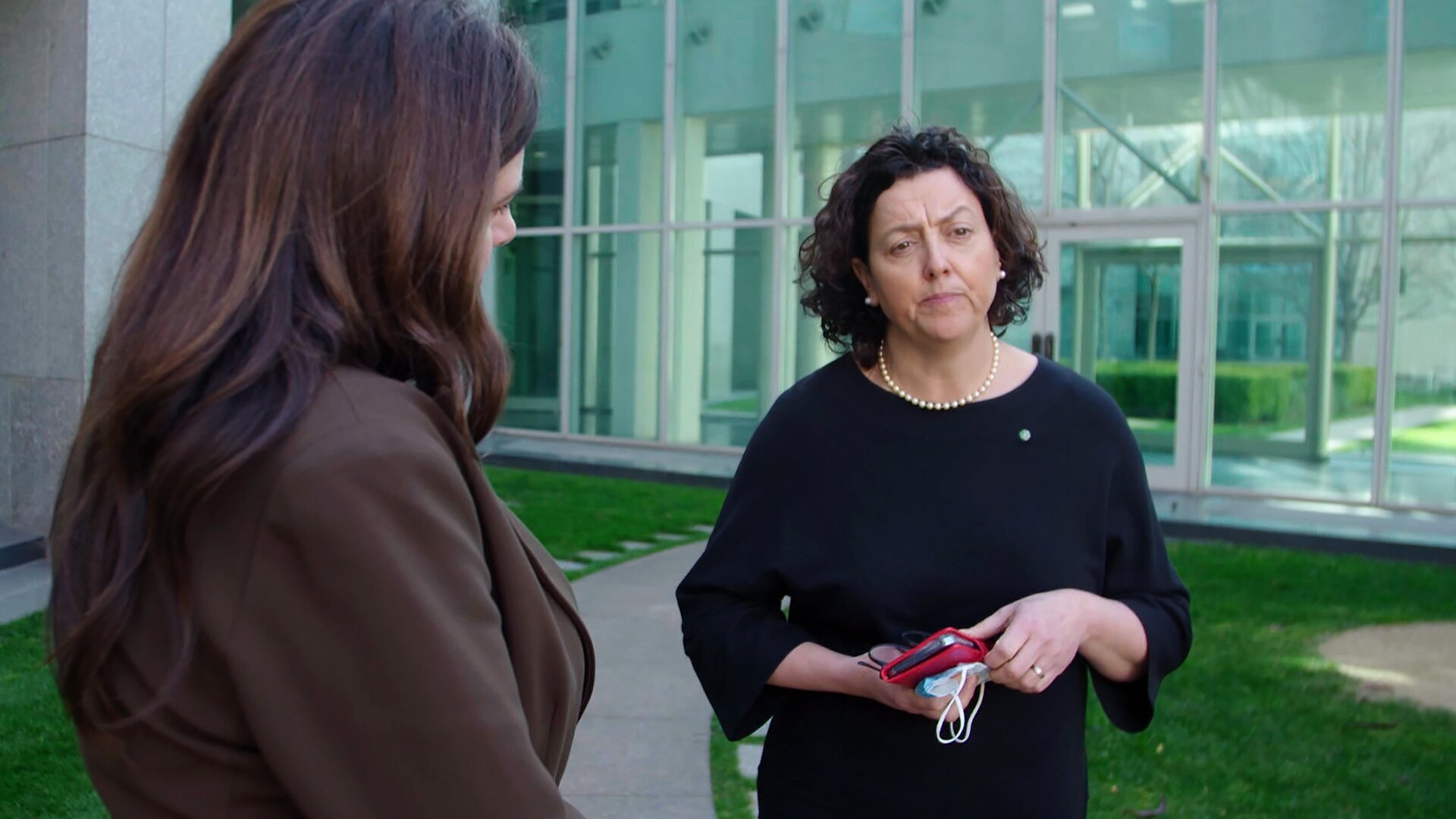 A woman in a black dress and pearls, carrying a mobile phone in a red case, chats with a journalist who is partly obscured