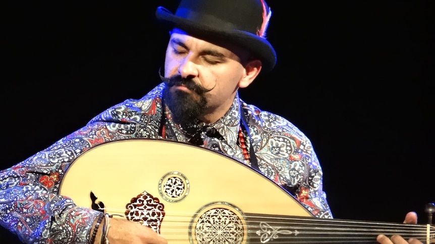 A man plays oud with his eyes closed. He wears a patterned shirt, bowler hat and beaded jewellery.