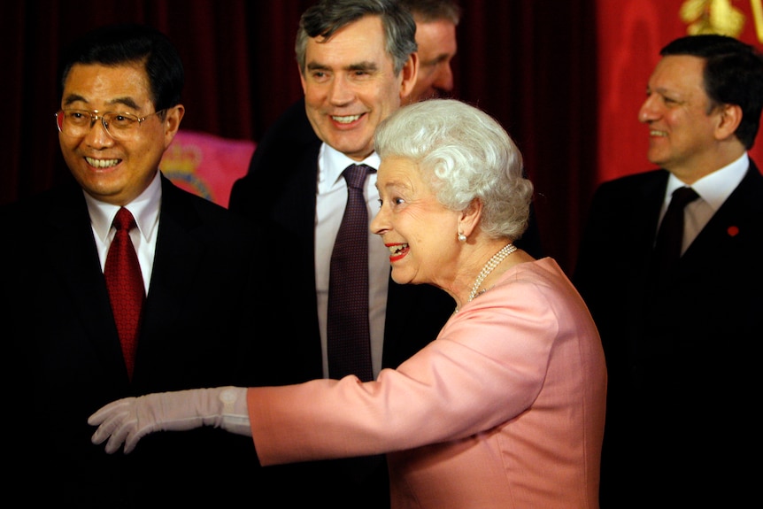 Queen in pink gestures with Gordon Brown and others smiling in background