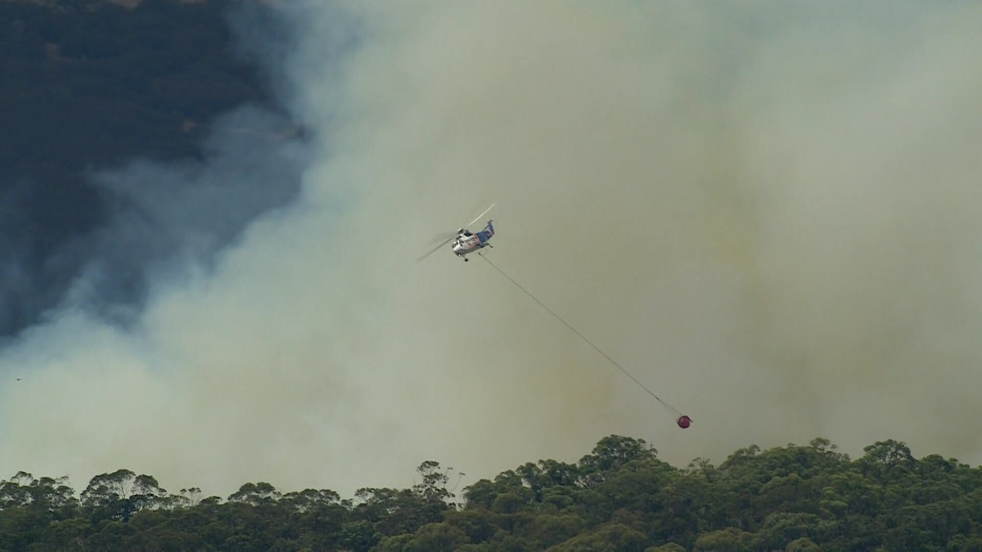 A helicopter with a water bucket hanging underneath it flies above green trees in front of white and brown smoke.