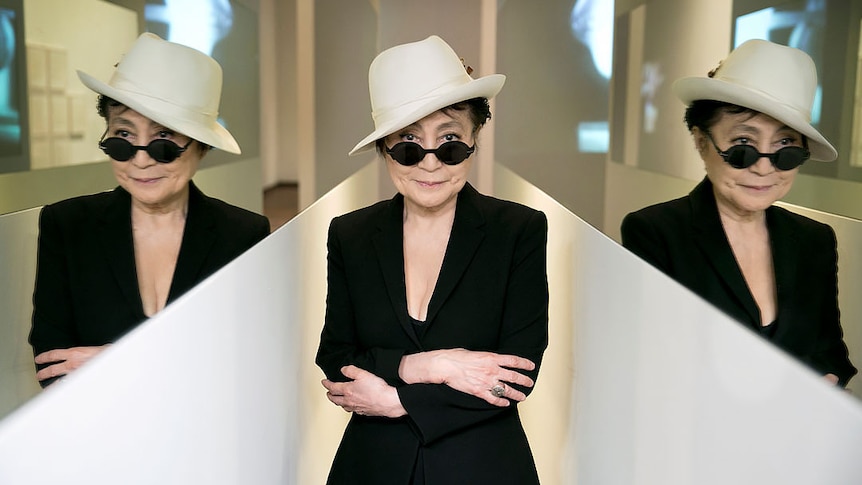 Yoko Ono stands in white hat, sunglasses, and black jacket in-between two mirrors showing her reflection