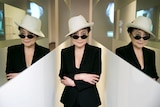 Yoko Ono stands in white hat, sunglasses, and black jacket in-between two mirrors showing her reflection