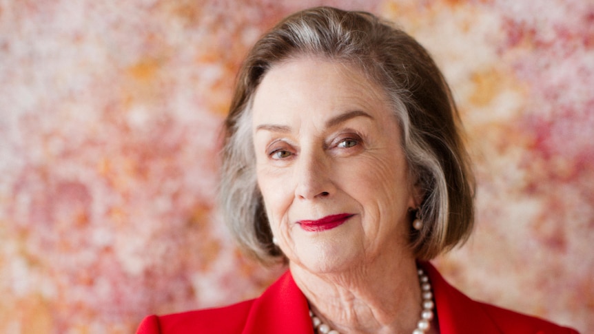 An elderly woman wearing a red blazer, smiles at the camera.