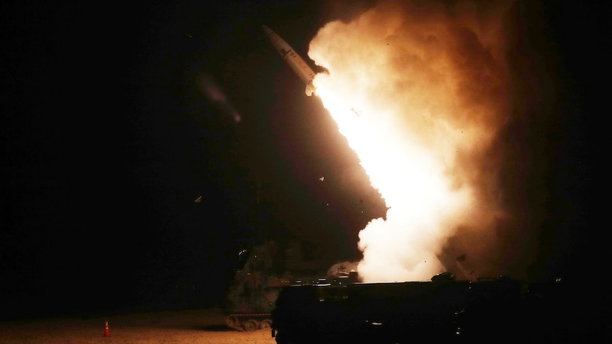 missile soars into the darkness with a plume of oranega nd white, tanks in foreground