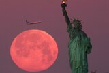 A pink coloured Super Snow Moon sets behind the Statue of Liberty as a plane passes by.