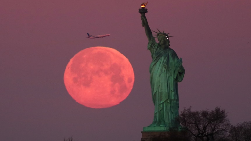 A pink coloured Super Snow Moon sets behind the Statue of Liberty as a plane passes by.