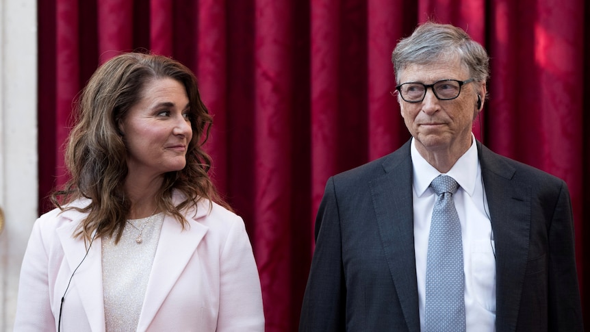 Melinda Gates smiles at Bill Gates while they stand in front of a red curtain. 