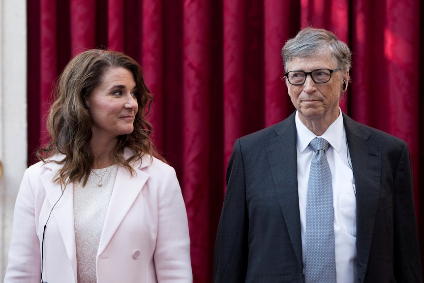 Melinda Gates smiles at Bill Gates while they stand in front of a red curtain. 