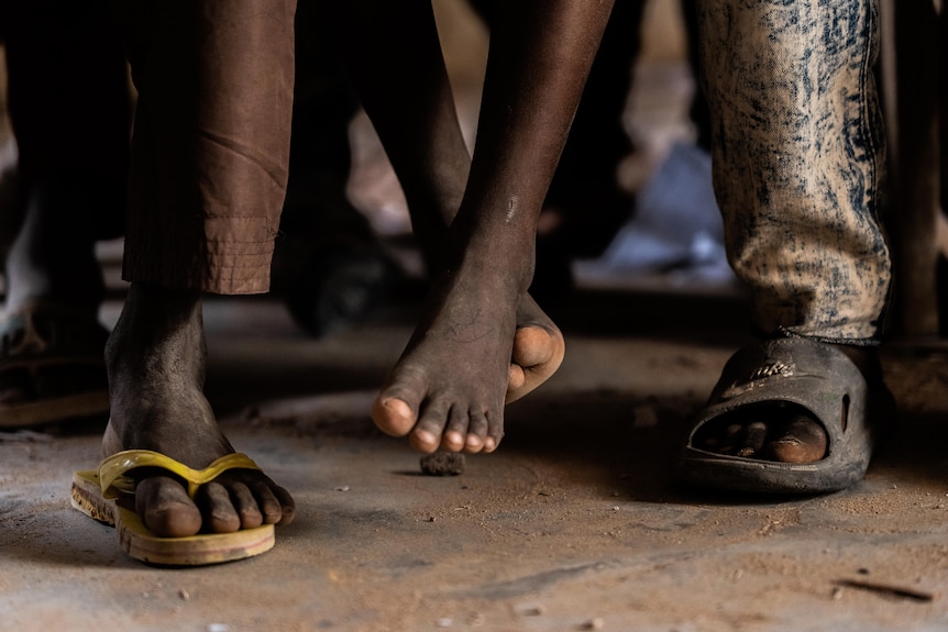 A close-up shows children's feet. One child is wearing no shoes, and their feet dangle above the dusty floor