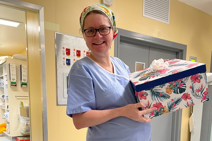 Katherine Goodall wears scrubs and holds a gift while pregnant