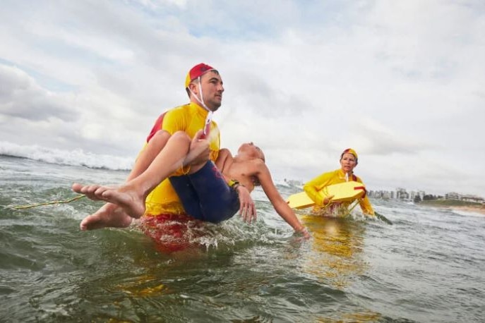 A man in lifesaving gear carries a limp child from the ocean as another lifesaver looks on.