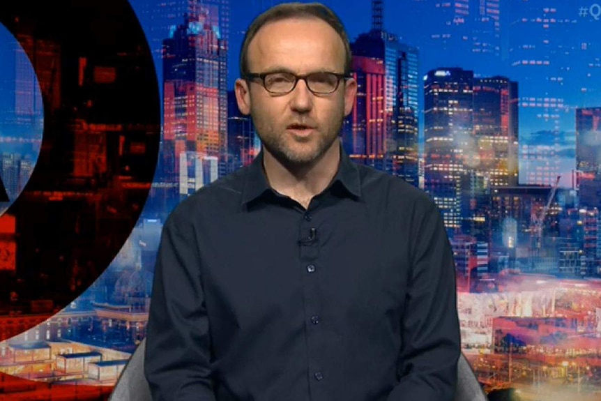 Adam Bandt is wearing glasses and a dark shirt set against a backdrop of Melbourne.