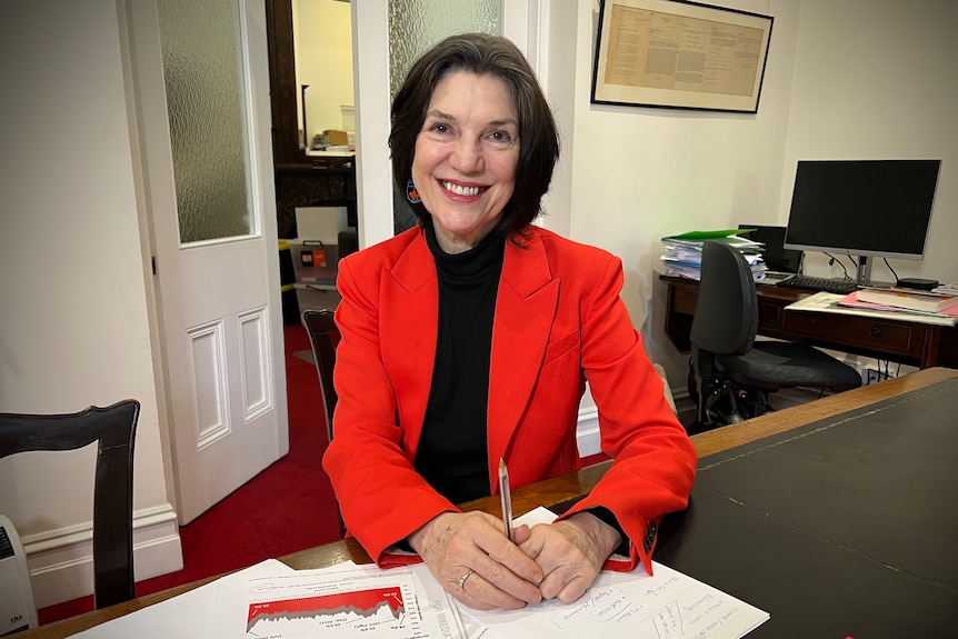 A woman in a red sports jacket and a black turtleneck sits at an office desk, smiling at the camera.