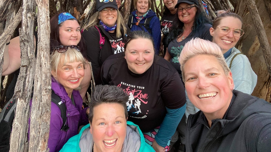 A group of 11 people in activewear smile widely as they crouch under a canopy of thin wooden sticks.