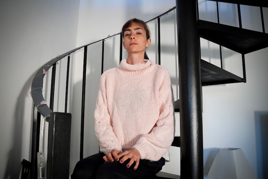 Sarah sits on a spiraling black staircase in her apartment, looking down seriously at the camera.