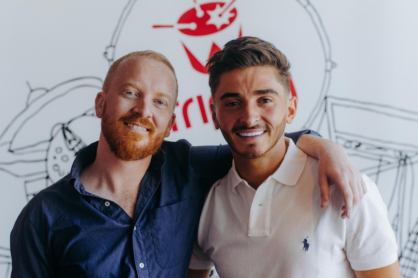 One man with red hair and a beard puts his arm around another young man with dark hair and a white polo shirt