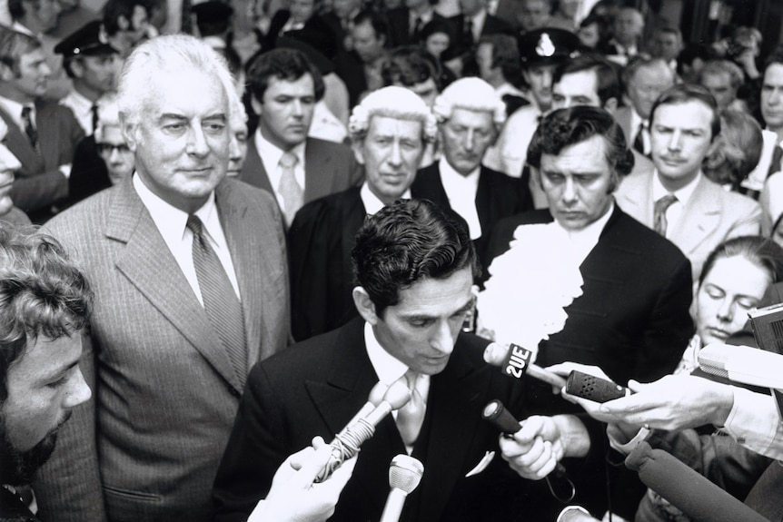 A man in a black suit reads from a piece of paper into some microphones.