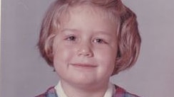 A aged school photo of a smiling primary school aged girl with short blond hair.