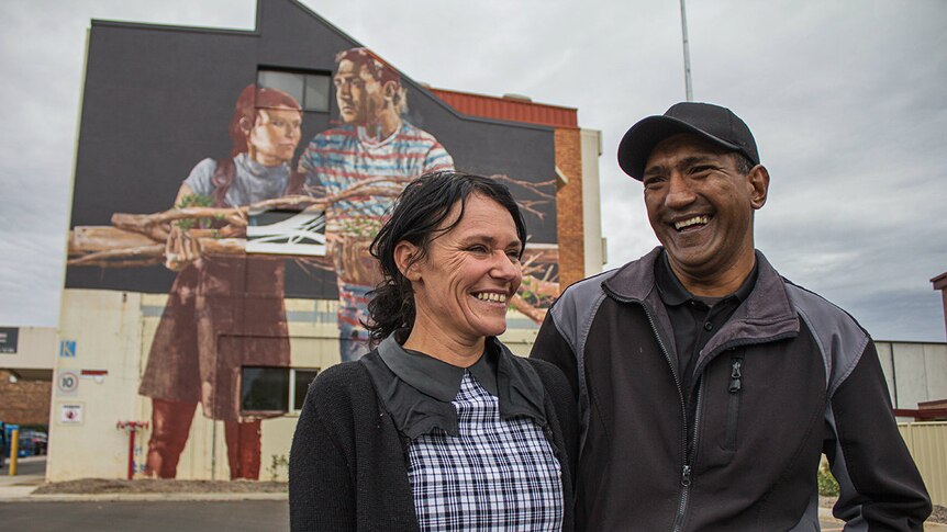 A couple stand smiling in front of a street mural