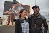 A couple stand smiling in front of a street mural