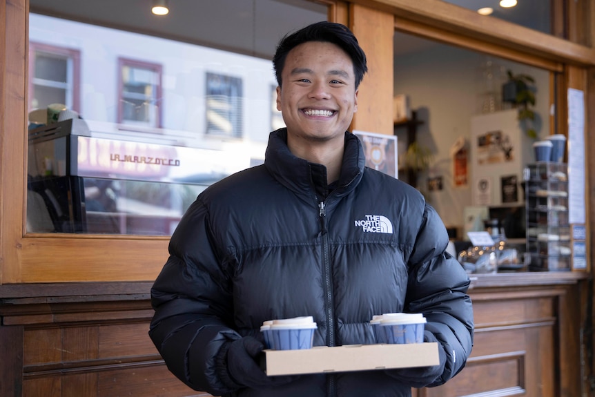 A man carrying two coffees in a tray smiles at the camera.
