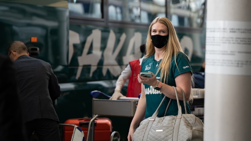 Ellen Roberts wearing a green jersey, holding a travel bag and wearing a black face mask, standing near a bus.