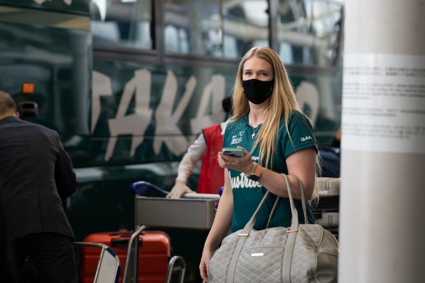 Ellen Roberts wearing a green jersey, holding a travel bag and wearing a black face mask, standing near a bus.