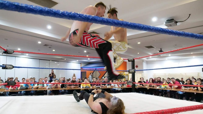 A crowd watches a wrestling match in a wrestling ring. Two men are in the air jumping over a man lying down in the ring.