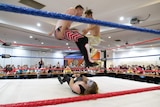 A crowd watches a wrestling match in a wrestling ring. Two men are in the air jumping over a man lying down in the ring.