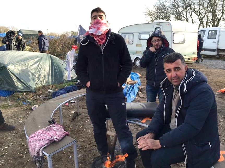 Amanj sits in the refugee camp warming his hands on a fire, surrounded by tents and rubbish covering the ground.