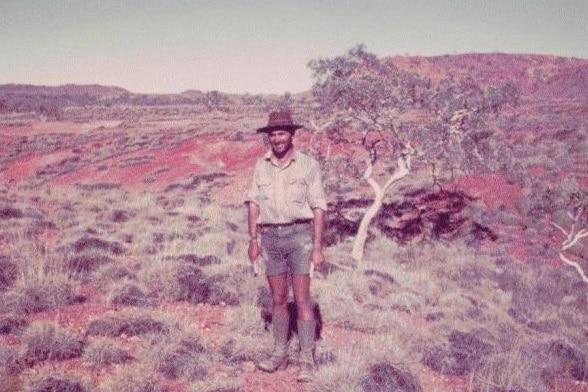 Jean-Paul Turcaud, wearing a hat, shorts, boots and big smile. standing in desert by scrubby tree.