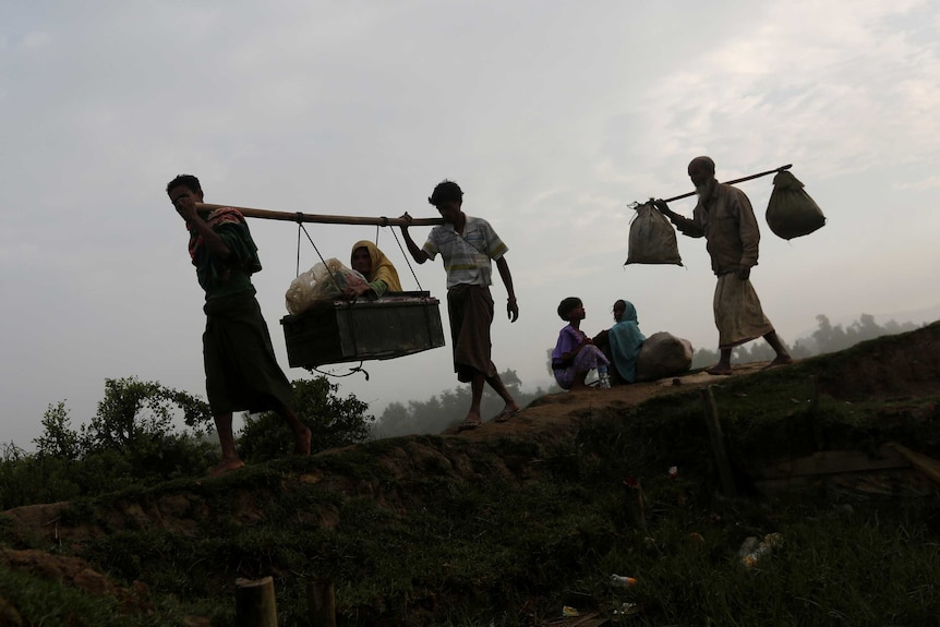 Two men carry an elderly woman in a basket and another man carries belongings.