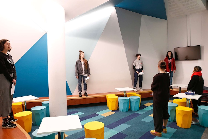 Four young performers stand on a bench in a university common room, performing to two creatives with their backs to the camera.