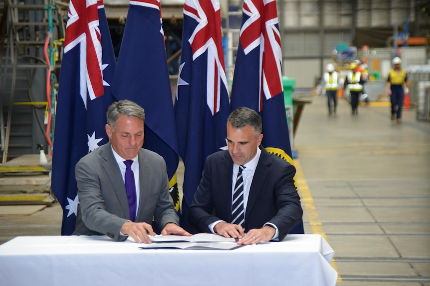 Two men sitting at a white table with papers in front of them. Behind them are Australian flags