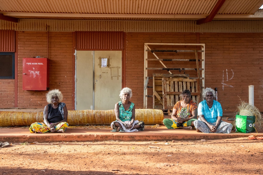 Four Burarra women in their 50s and 60s sit on concrete in the afternoon sun in front of a large weaving rolled up.
