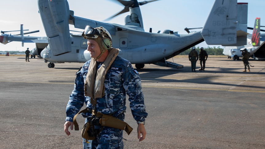 A man in fatigues and a pilot's cap walks along the tarmac, a large military plane behind him.